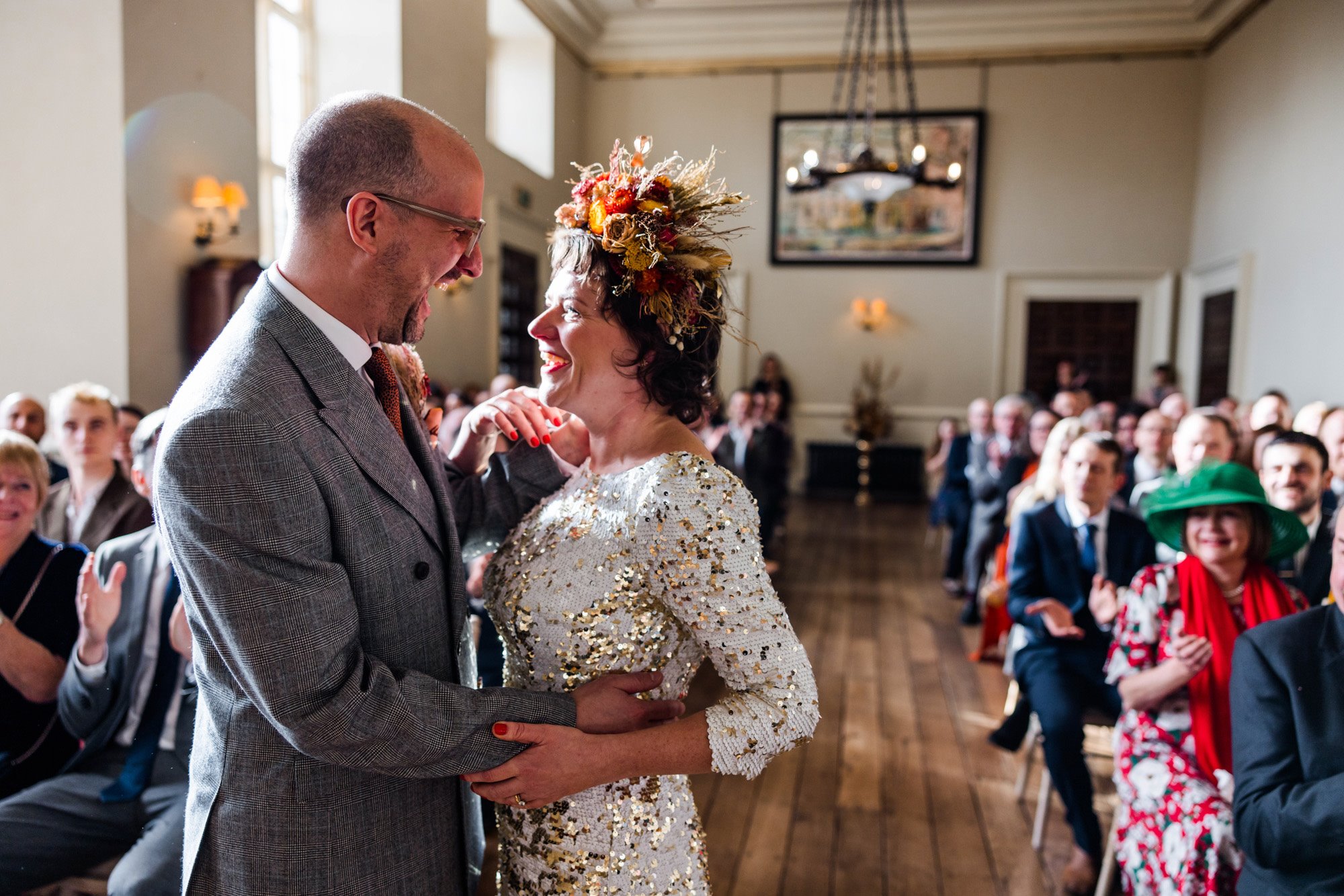 Older bride in flower headdress and sequin dress and groom in grey suit in joyful embrace at wedding ceremony in stately home