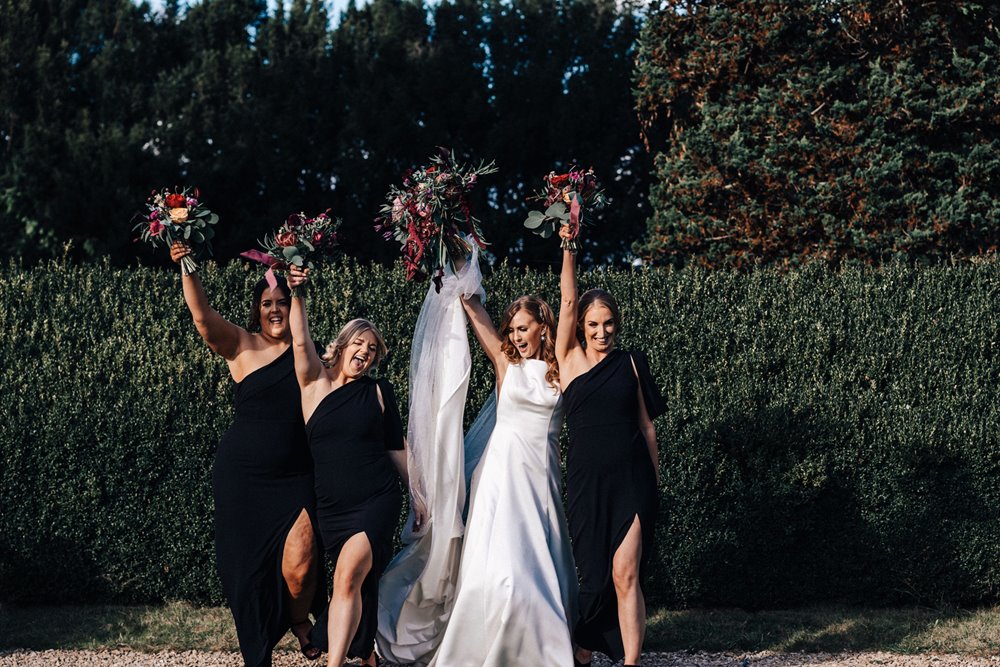 Cool bride and bridal party in black bridesmaids dresses holding up autumnal bouquets at dark and moody fun wedding in October
