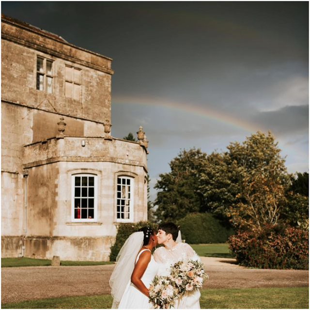 Two brides kiss under rainbow over mansion house on their beautiful gay wedding day at elmore court in gloucestershire 
