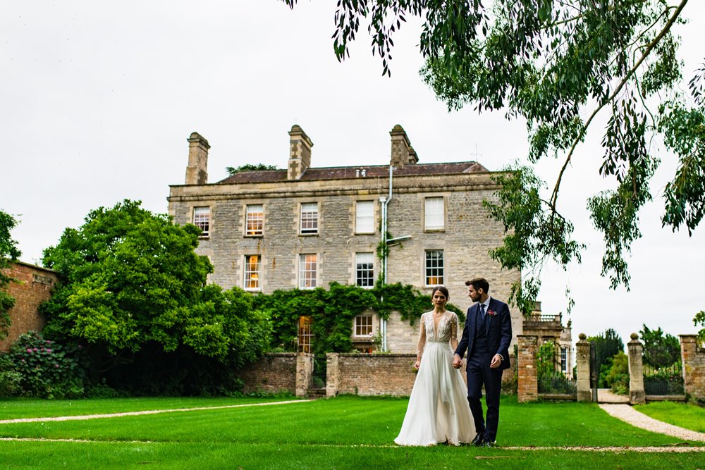 elegant bride and groom walk through the walled garden hand in hand with stately home elmore court in the background