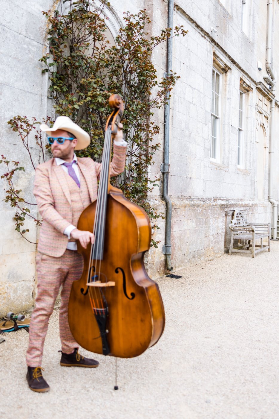 Half of the cash cows musical duo man playing double bass in pink suit and cowboy hat and blue plastic sunglasses in front of mansion house wedding venue elmore court