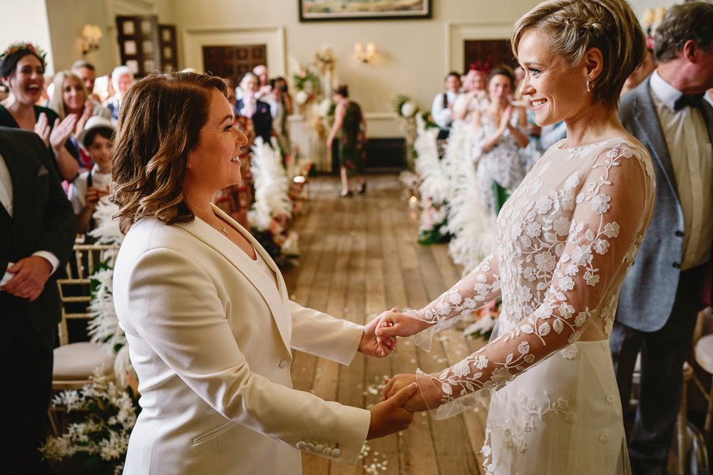Two brides holding hands facing each other at beautiful wedding ceremony in stately home venue