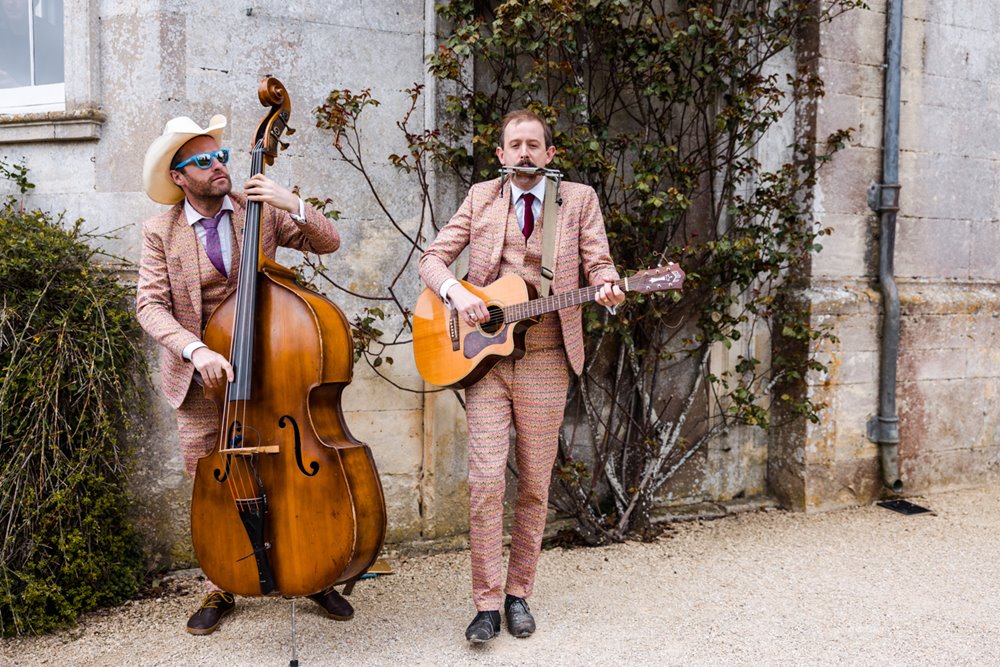 The cash cows musical duo dressed in pink and brown suits and cowboy hat play double bass and guitar and harmonica outside elmore court mansion house wedding venue in gloucestershire