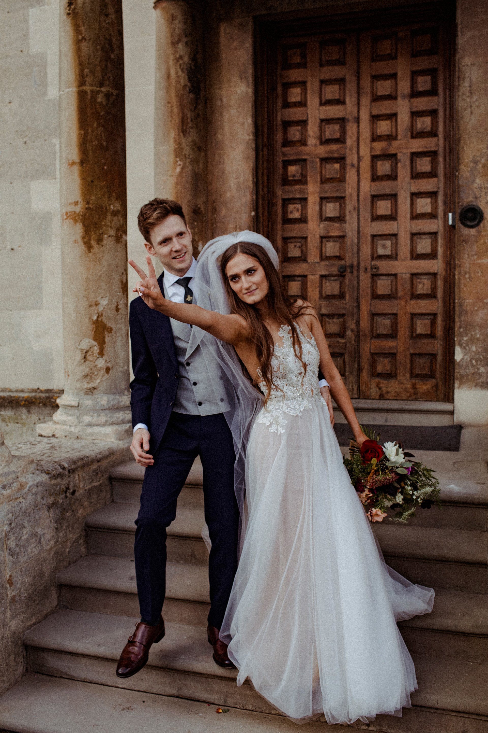 Cool Bride pulls v sign pose with groom outside their mansion house autumn wedding venue