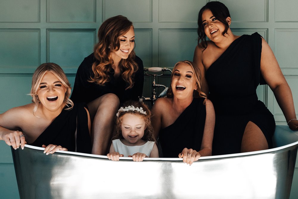 bridesmaids wearing black dresses laugh and pose in huge silver bath tub on the morning of wedding in a mansion house in the cotswolds