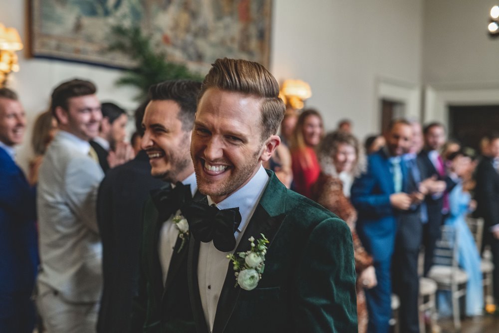 Handsome groom laughs at the camera walking down the aisle with his new husband wearing matching tuxedos at same sex ceremony