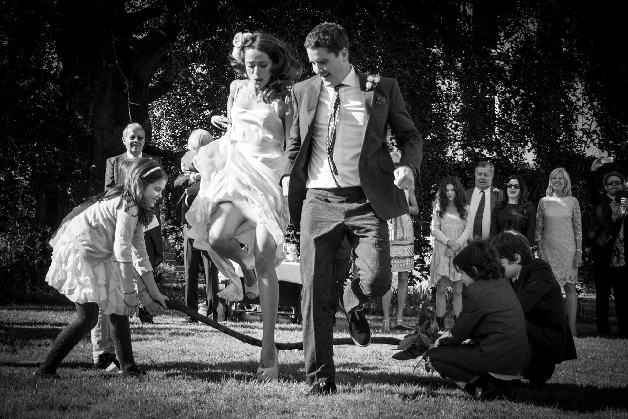 Jumping the broom ceremony- bride and groom jump over a stick held by two children on the lawn outside their festival wedding venue elmore court