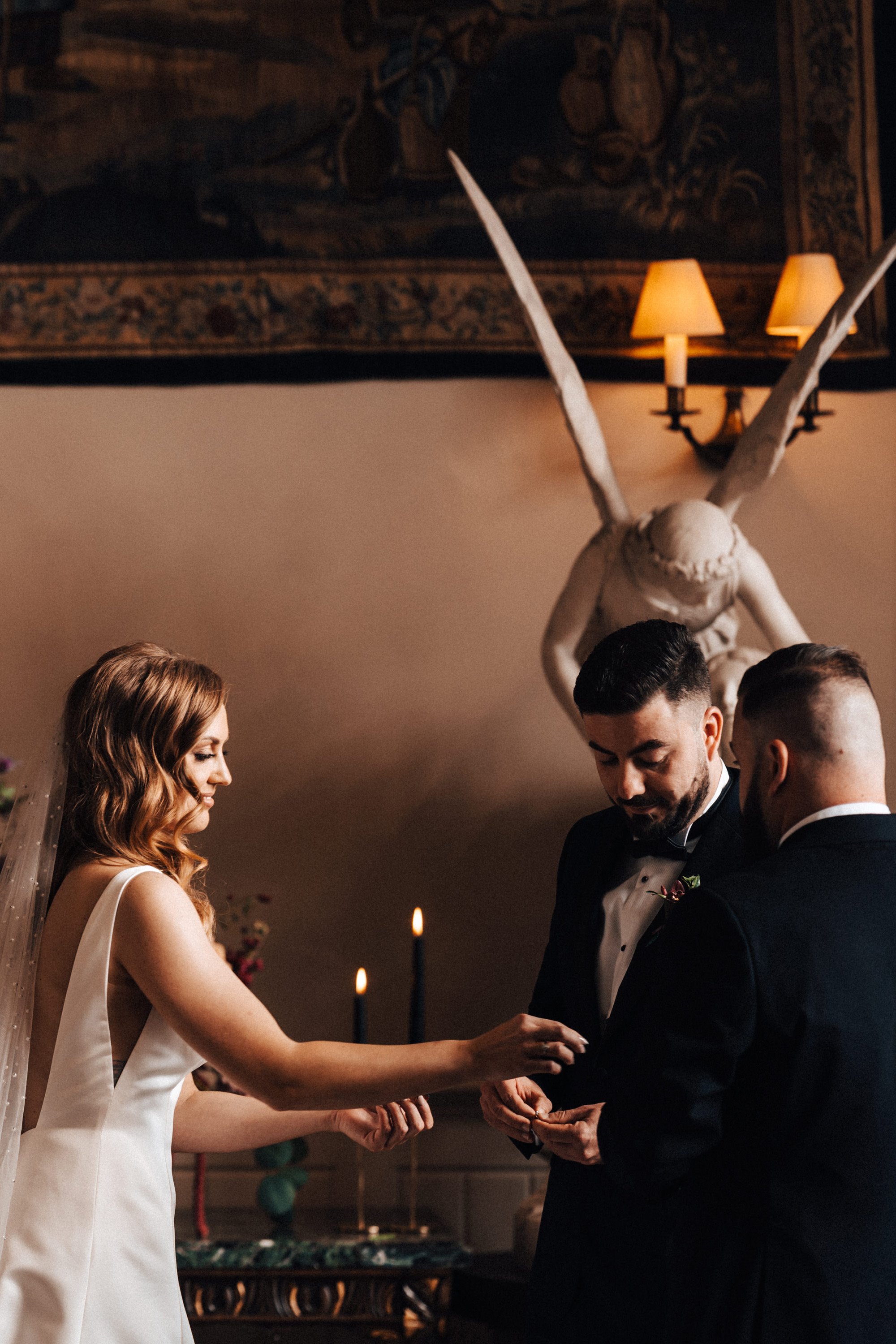 bride and groom exchange rings at Moody october wedding ceremony with black candles and angel statues in historic country house in gloucestershire