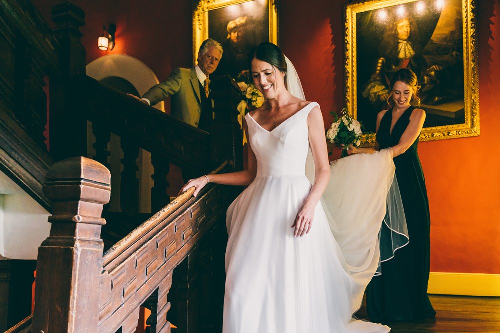 Bride descends down the stairs of stately home with guests carrying her train ready to marry at a micro wedding in 2020