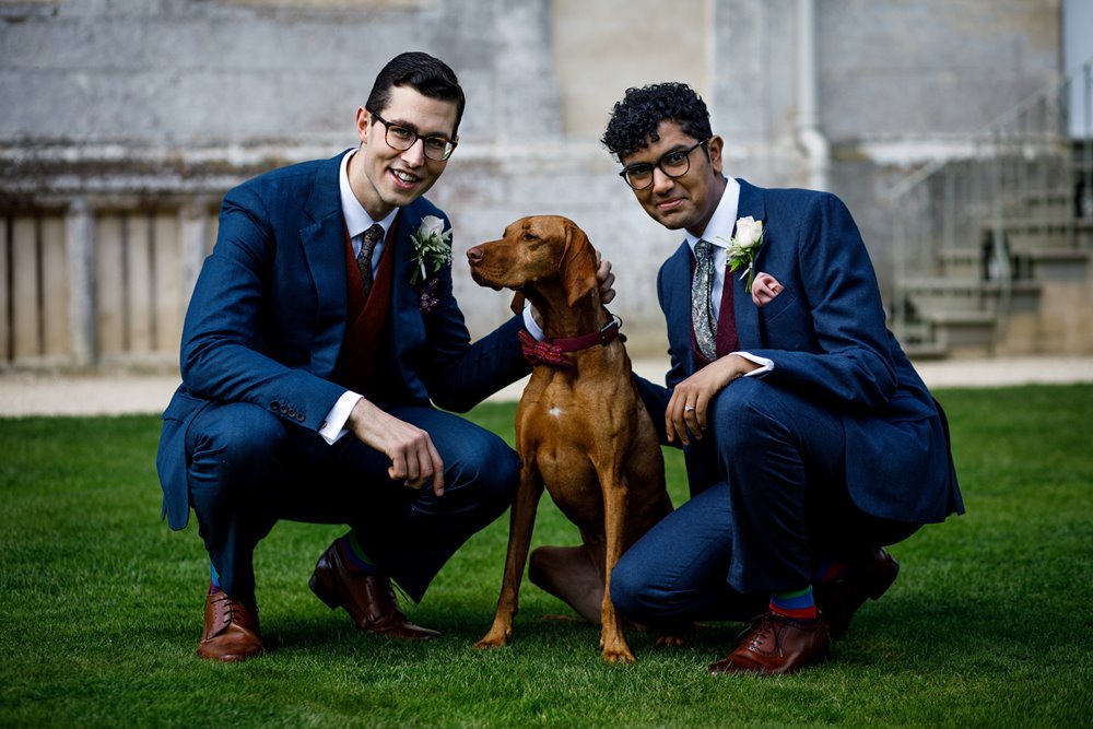 Two grooms and a dog page boy smile outside their stately home wedding venue