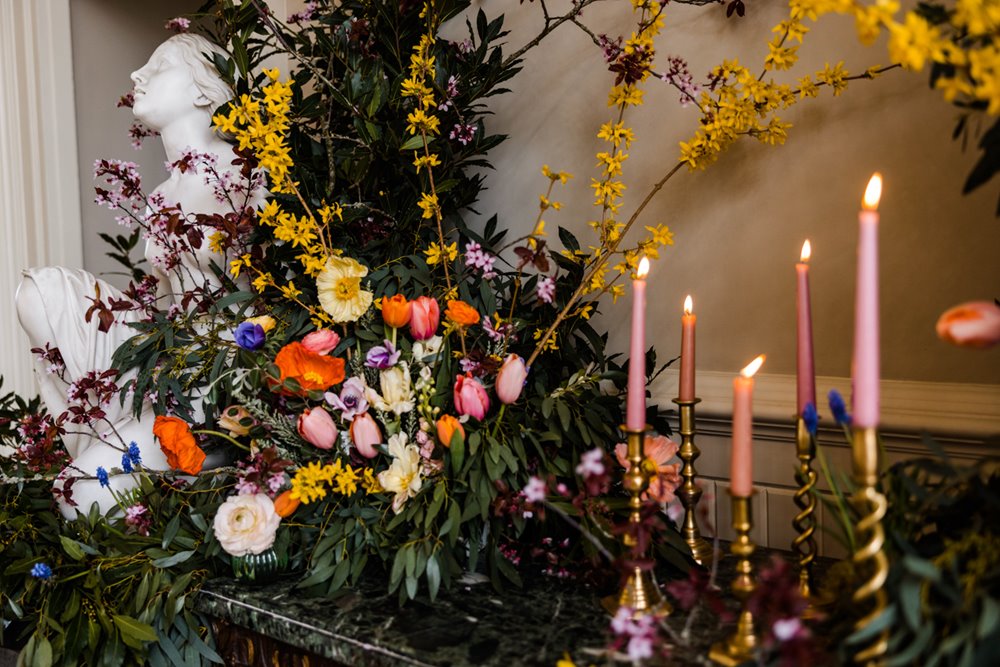 Pink tapered candlesticks with wild wedding flowers decorate wedding ceremony hall with huge stone statue of winged woman at mansion house wedding venue in gloucestershire