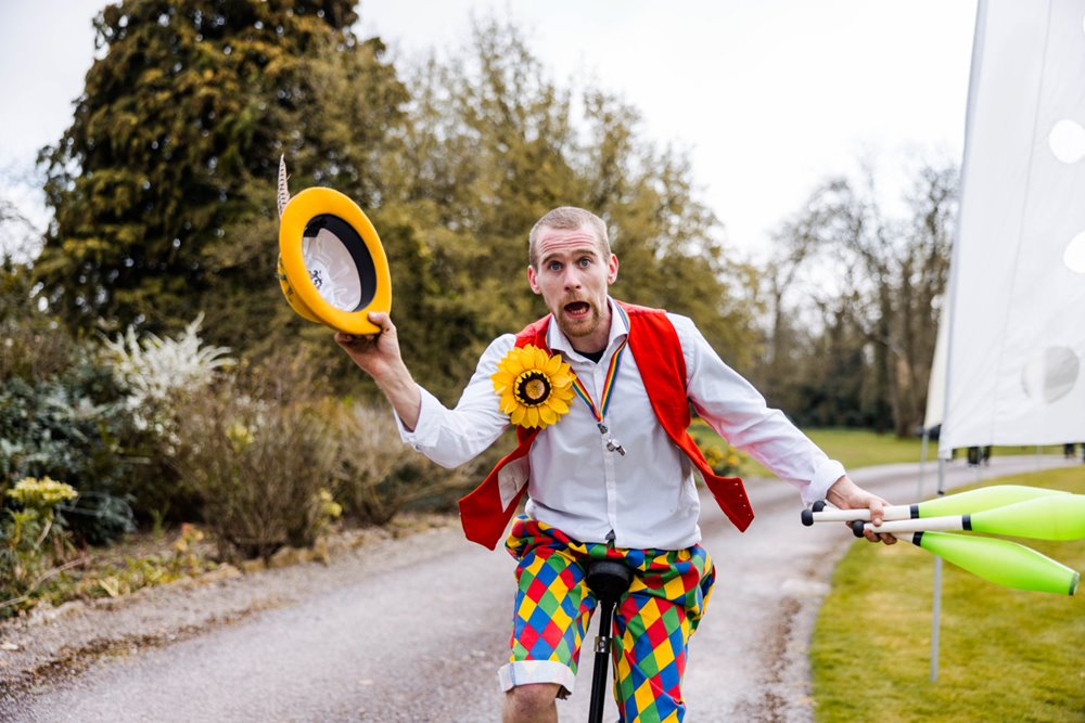 Circus entertainer on unicycle lunges towards the camera looking worried with yellow hat in his hand at unusual wedding fair open day in the cotswolds