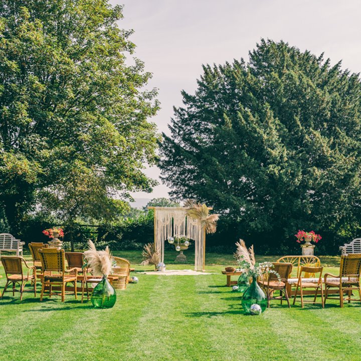 Boho outdoor wedding ceremony with macrame arch mismatching chairs and pampas grass in glass pots on the lawn at elmore court in the cotswolds