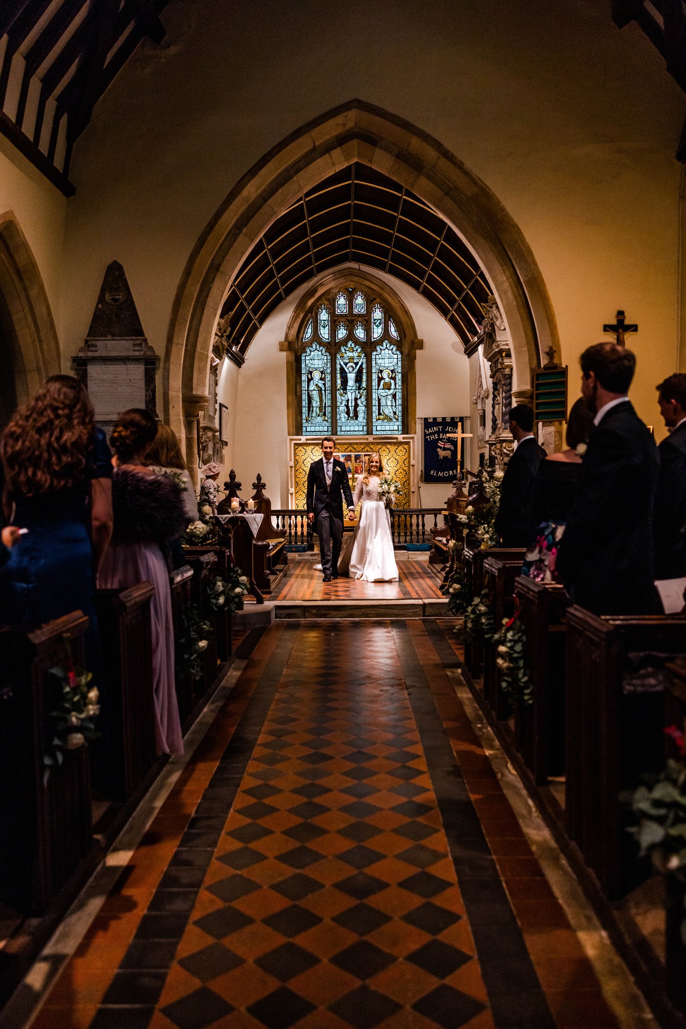 Pretty church wedding bride and groom walk towards with stained glass window in background and atmospheric dim light