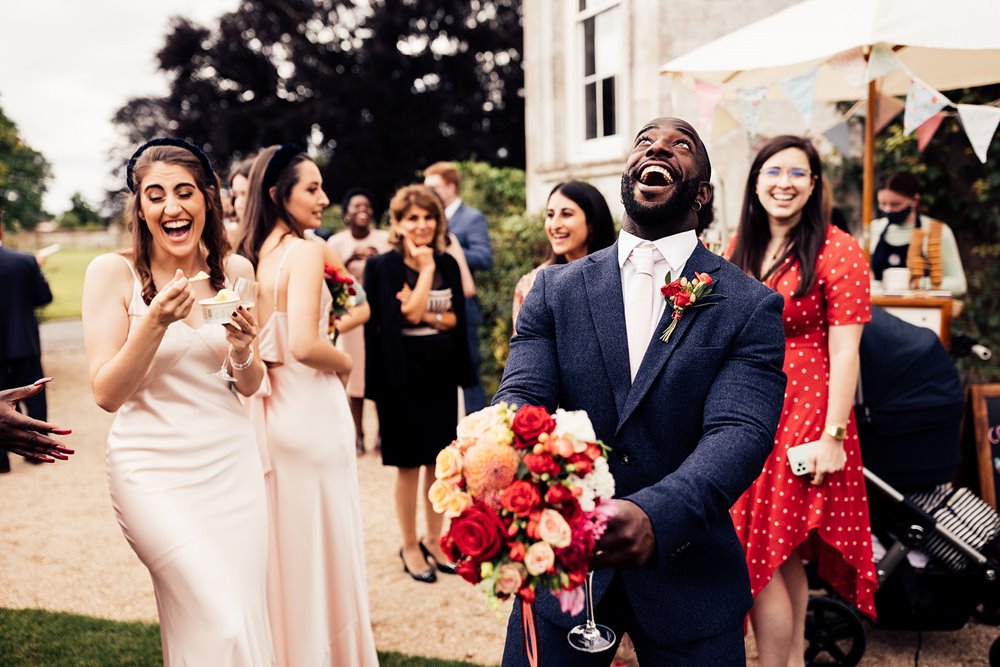 Bridesman throws wedding bouquet guests laugh outside stately home weddign venue elmore court in the costwolds 