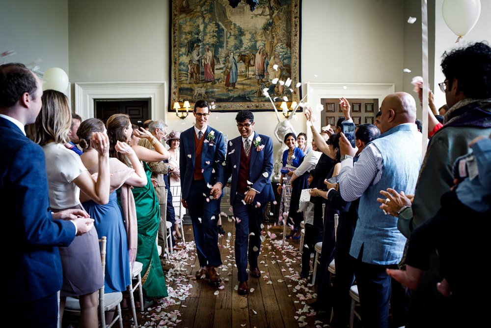 Two grooms walk happily down the aisle surrounded by guests throwing petal confetti