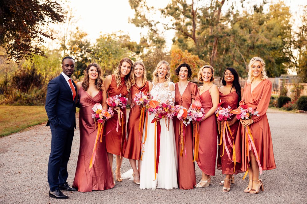 bridesman and bridesmaids in burnt orange copper dresses stand with boho bride with bright bouquets outside stately home wedding venue in the cotswolds countryside 