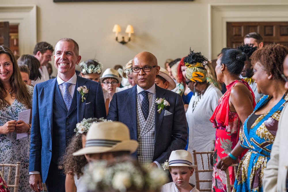 Gay wedding couple enter their ceremony in the hall surrounded by guests at Elmore Court