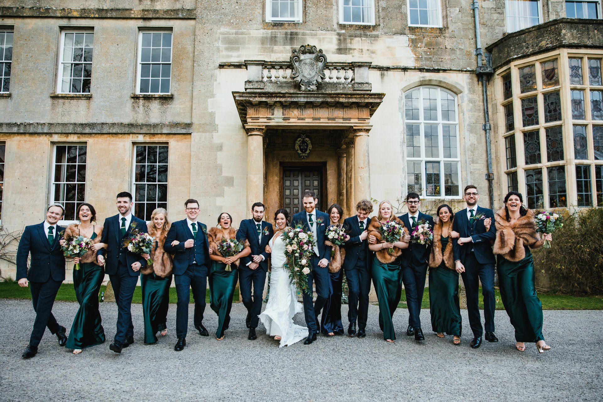 Big wedding Bridal party and groomsmen link arms and laugh in front of their weekend wedding venue