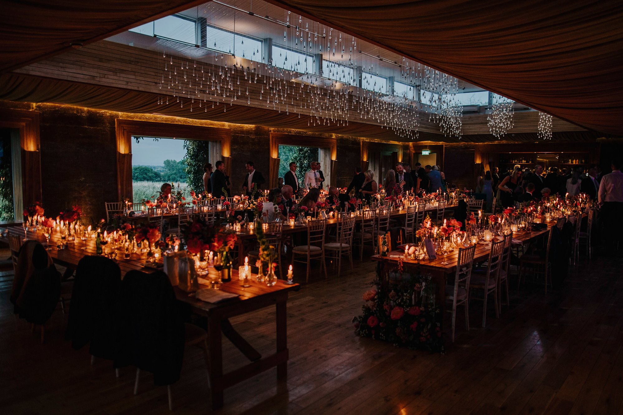 Guests enjoying a wedding reception in a candle lit Gillyflower at Elmore Court, with tables dressed in pink flowers, candles and place settings
