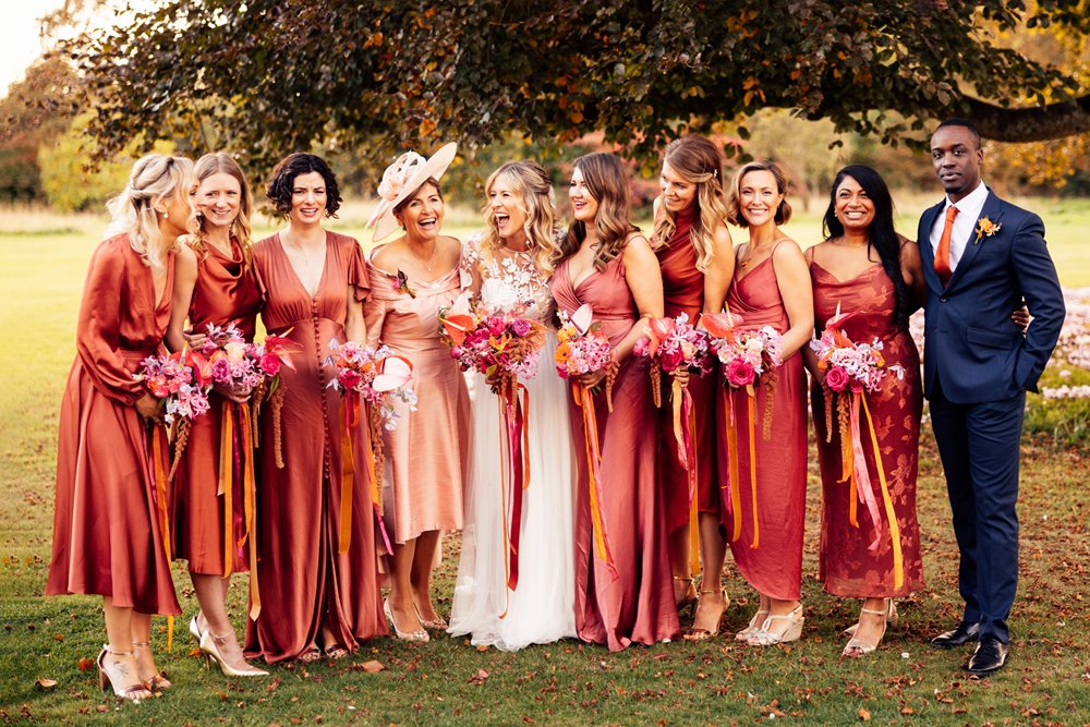 Colourful bridesmaids in orange dresses stand with boho bride with bright bouquets with ribbons in the cotswolds countryside 