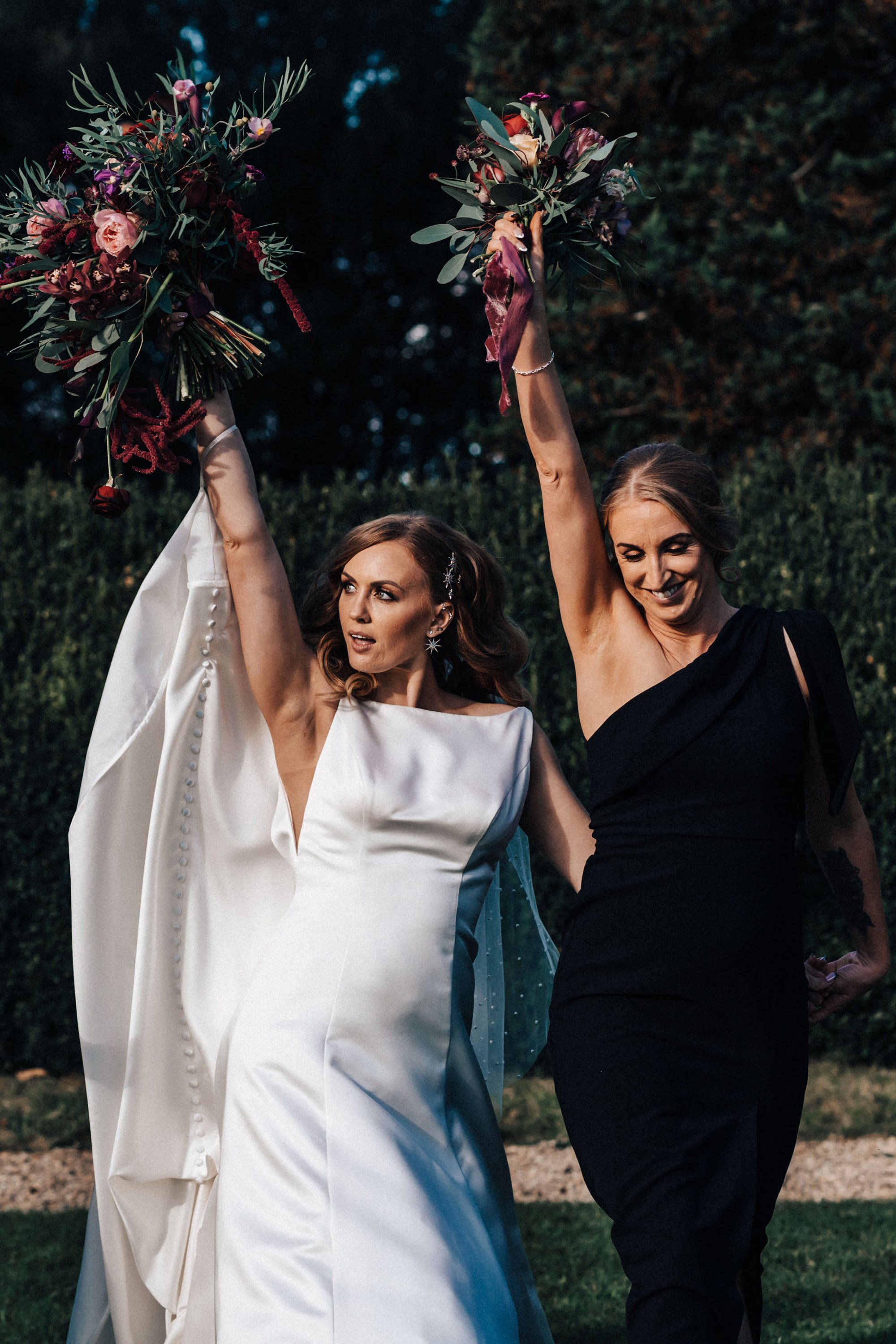 Bride poses with maid of honor in black bridesmaids dress holding up dark autumnal wedding flowers at Elmore Court in Gloucestershire