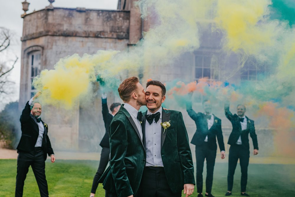 Rainbow smoke bombs go off behind gay couple kissing in green velvet suits on their wedding day