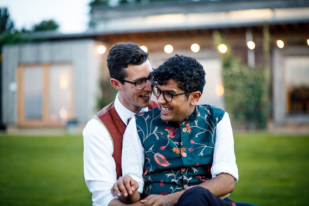 Gay couple laugh holding each other with lights in background outside their wedding venue