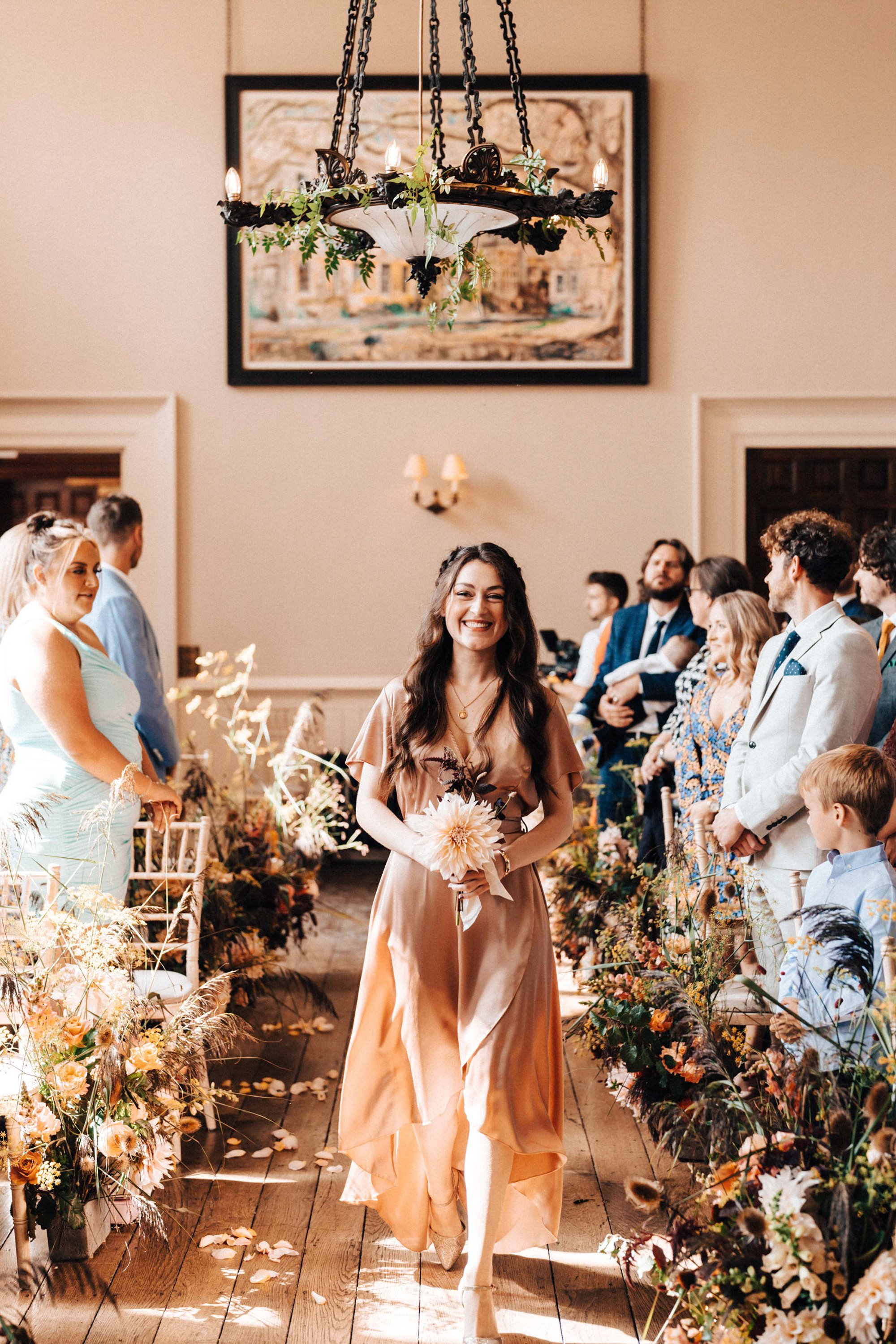 Bridesmaid in pink silk dresses walks down a floral aisle in beautiful mansion house wedding venue