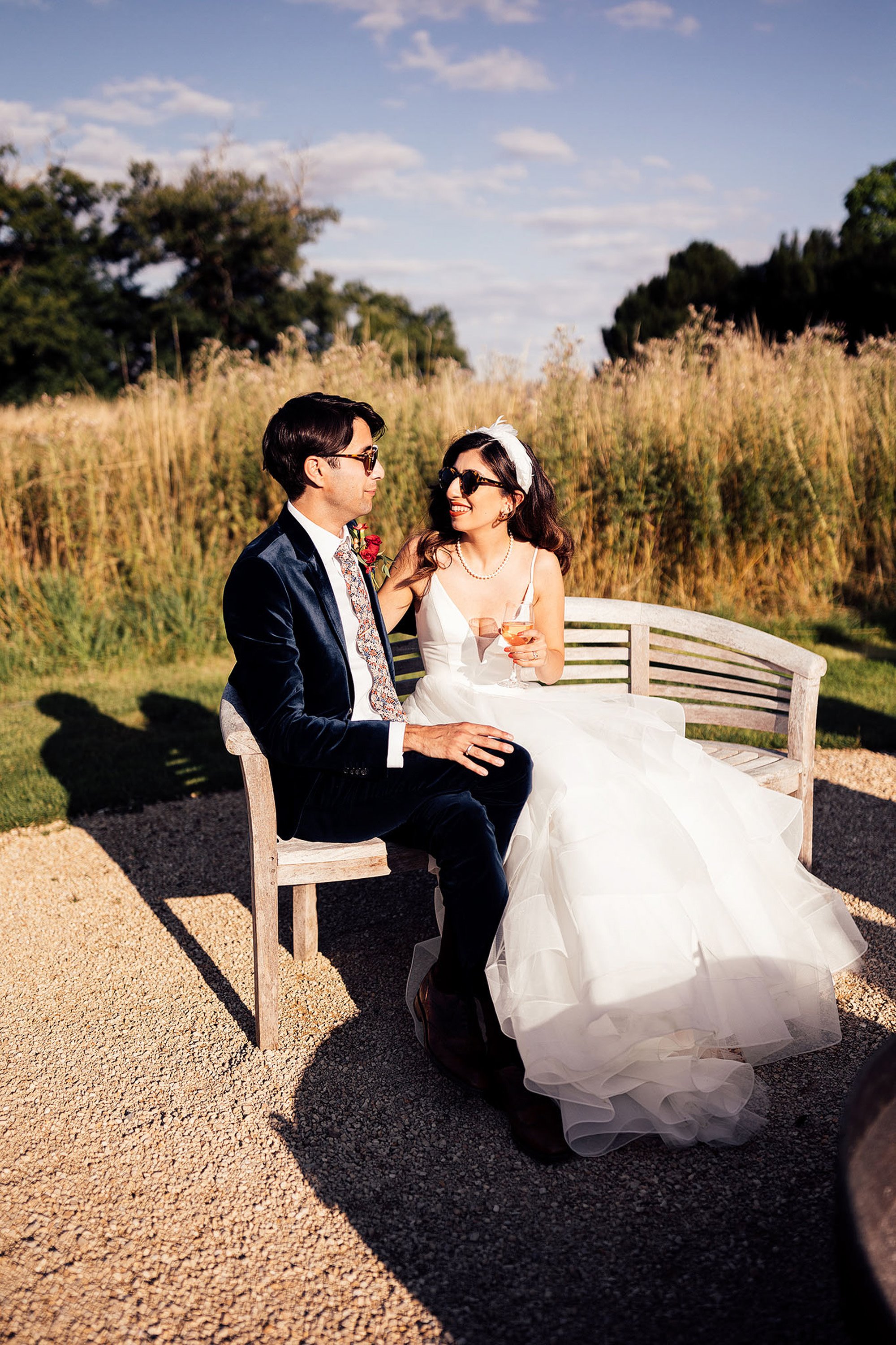 Gorgeous Persian Cool bride and groom sit on bench in meadow in sunshine wearing sunglasses. Bride wears pearls, headpiece and full dress and red lipstick, groom wears velvet suits and floral tie