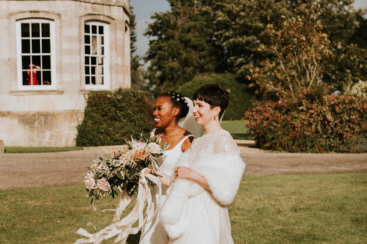 Two brides in flowing long white wedding dresses and fur wrap smiling standing side by side underneath a rainbow in the sky above their stately home wedding venue on their same sex ceremony in the cotswolds countryside