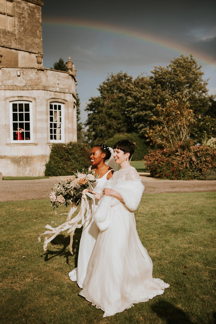 Two brides in flowing long white wedding dresses and fur wrap smiling standing side by side underneath a rainbow in the sky above their stately home wedding venue on their same sex ceremony in the cotswolds countryside