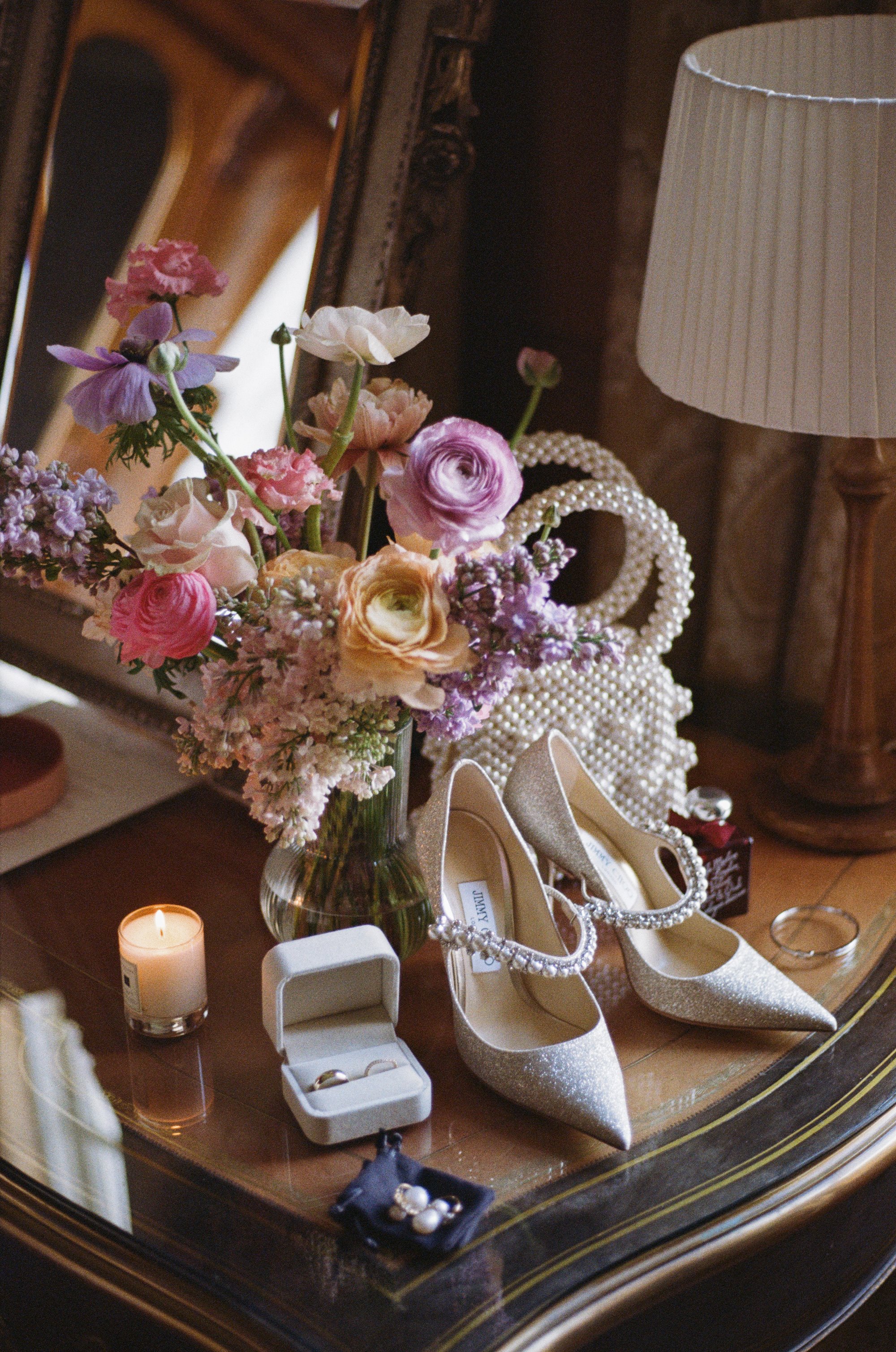 stunning wedding accessories on an antique table along with pearl adorned heels, wedding rings, handbags and a bouquet of flowers