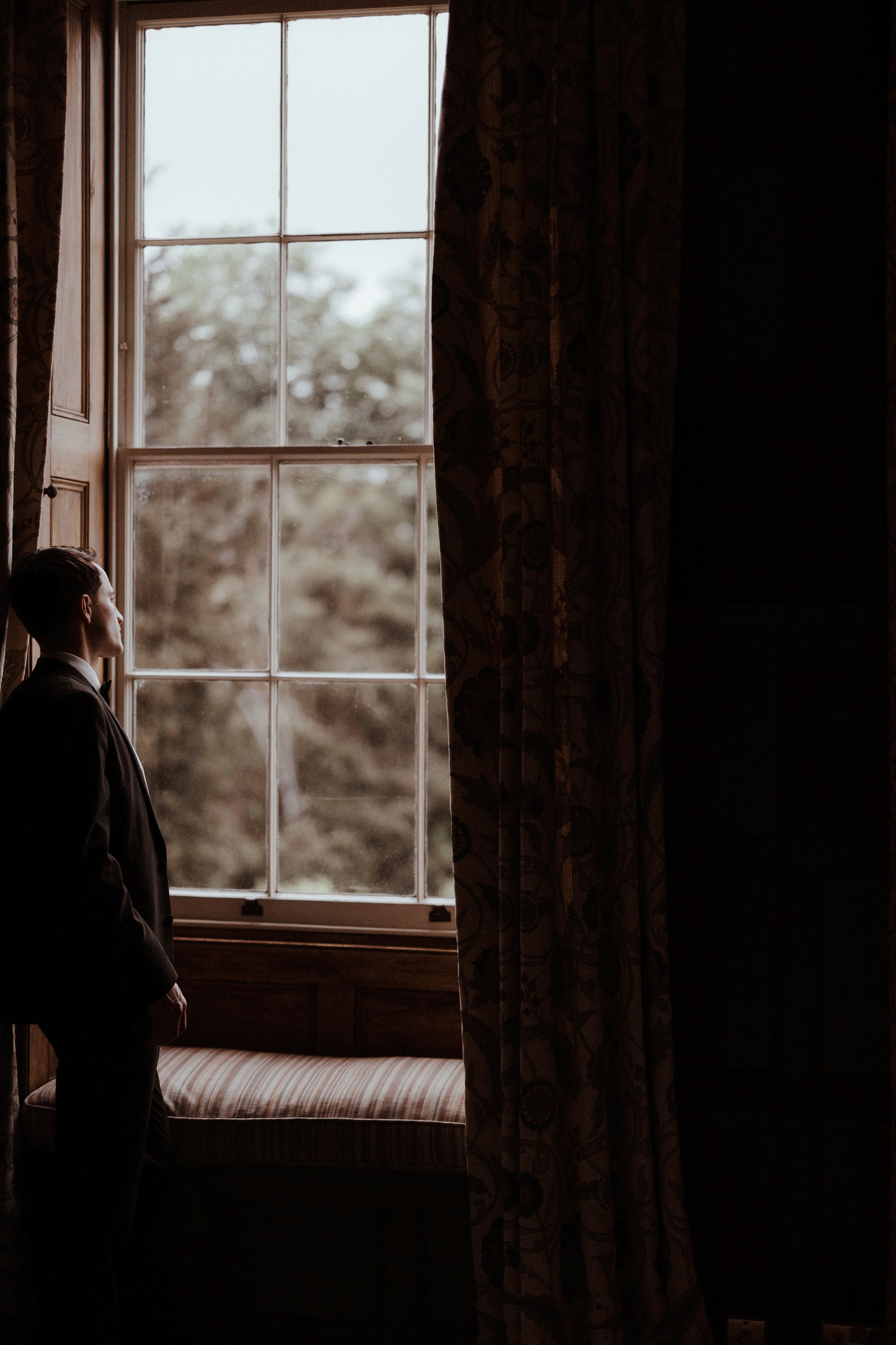 Fine art wedding photo of groom looking out window of stately home on wedding day in the costwolds