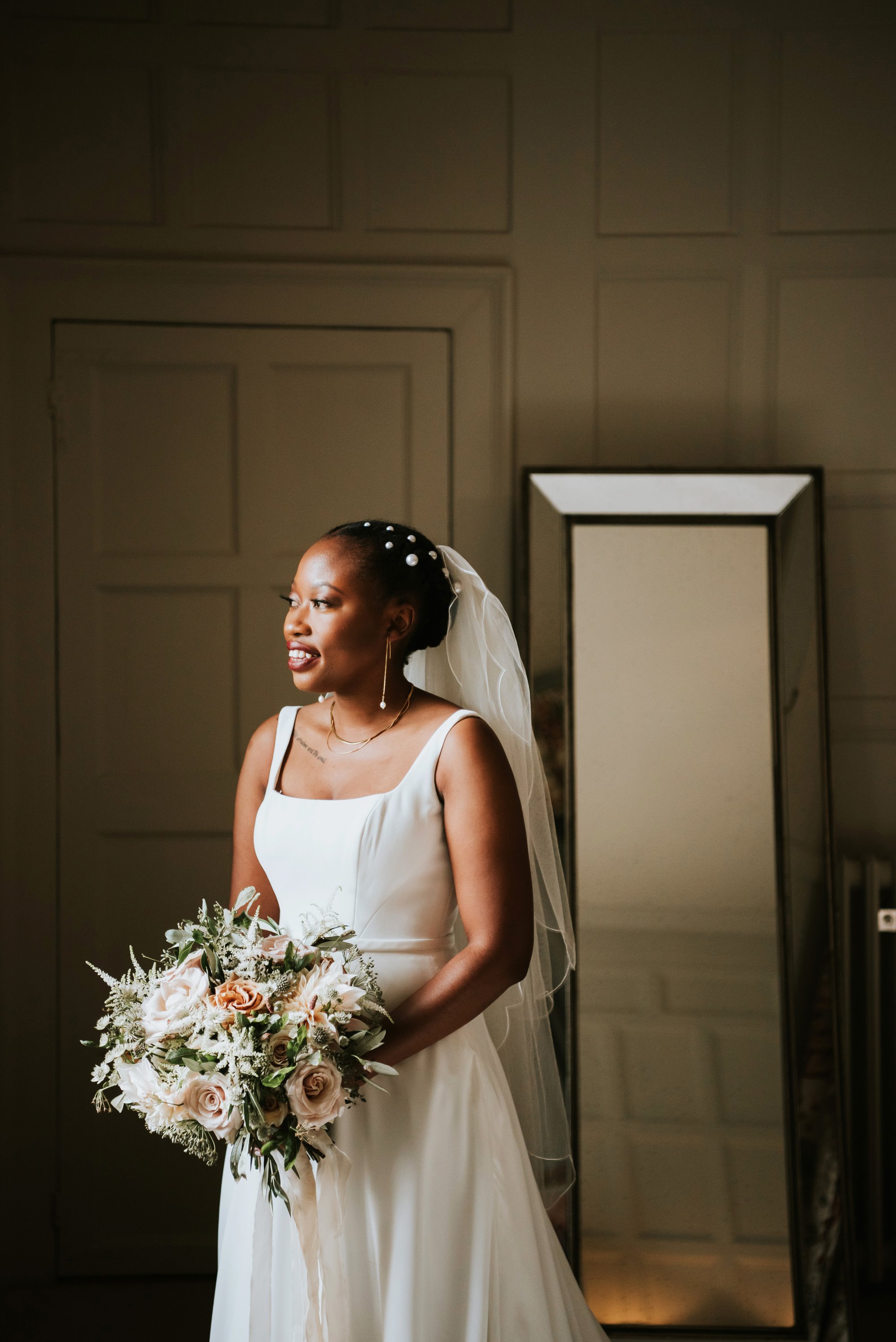 Modern black bride wearing simple satin dress with pearl earrings and pearl hair pins holding chic bouquet of pink roses and greenery