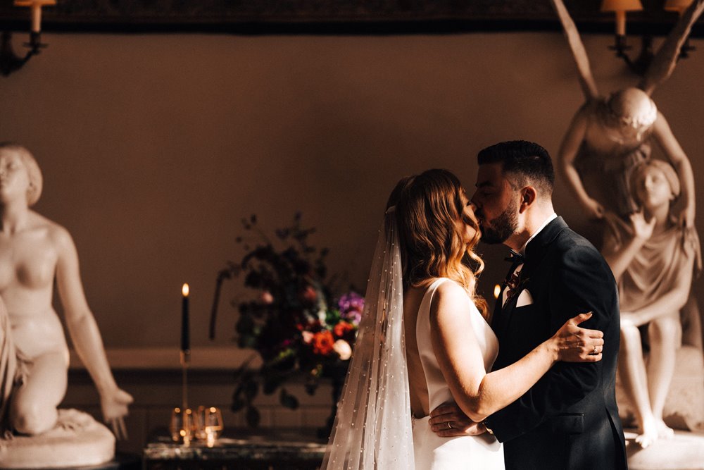 Bride and groom kiss in front of black candles and angel statues at their Moody October wedding ceremony in an old mansion house in England