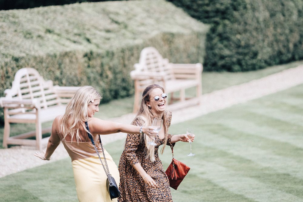 Two female wedding guests dance and laugh on the lawn at a summer wedding at Elmore Court