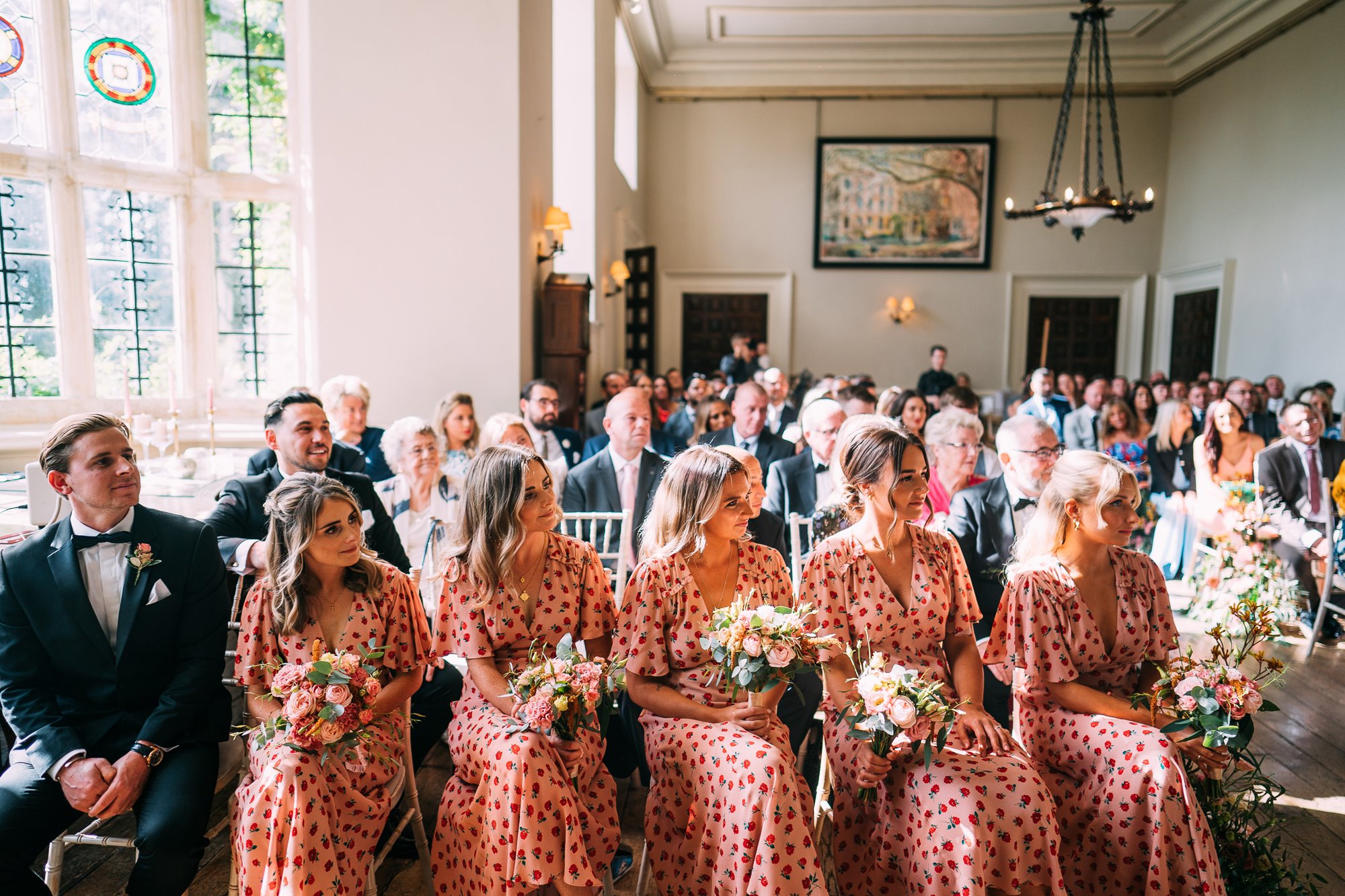 Bridesmaids sitting in front row of wedding ceremony in pink and red dresses holding pink bouquets at Elmore Court