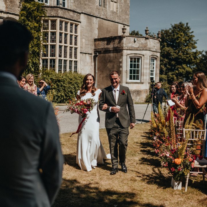 Gorgeous bride and groom walking across a lawn, in front of a stately home, for their beautiful outdoor wedding at Elmore Court 