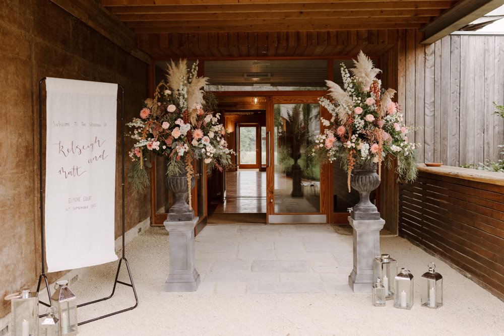 Two urns full of big floral displays with pampas and soft pink flowers outside an eco wedding reception venue in Gloucestershire
