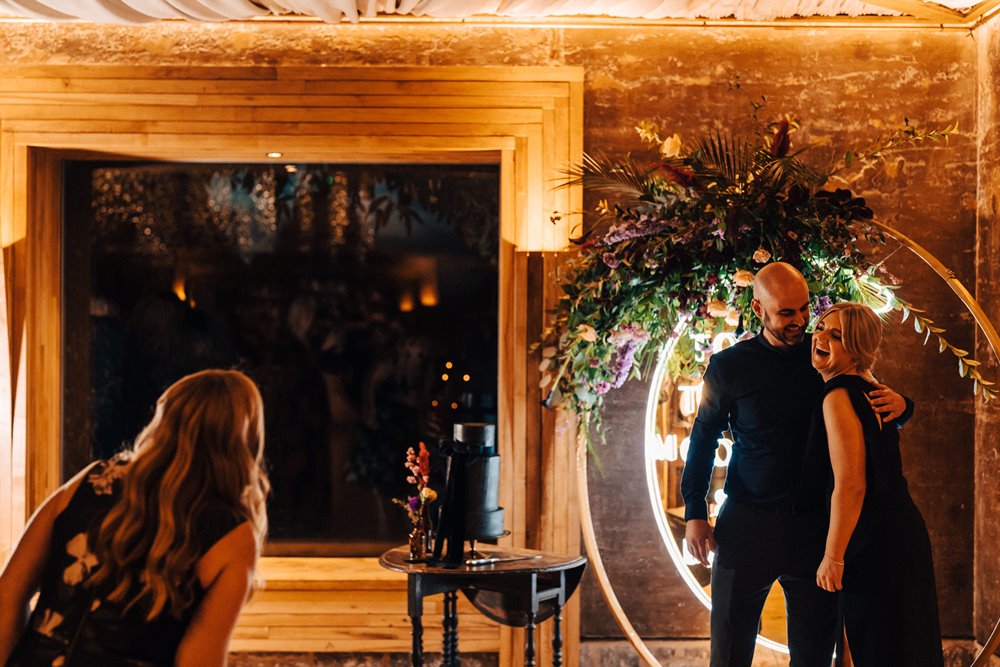 Wedding guests and bridesmaids wearing black posing in front of light up crescent moon decorated with autumn flowers for a moody October wedding with black accents