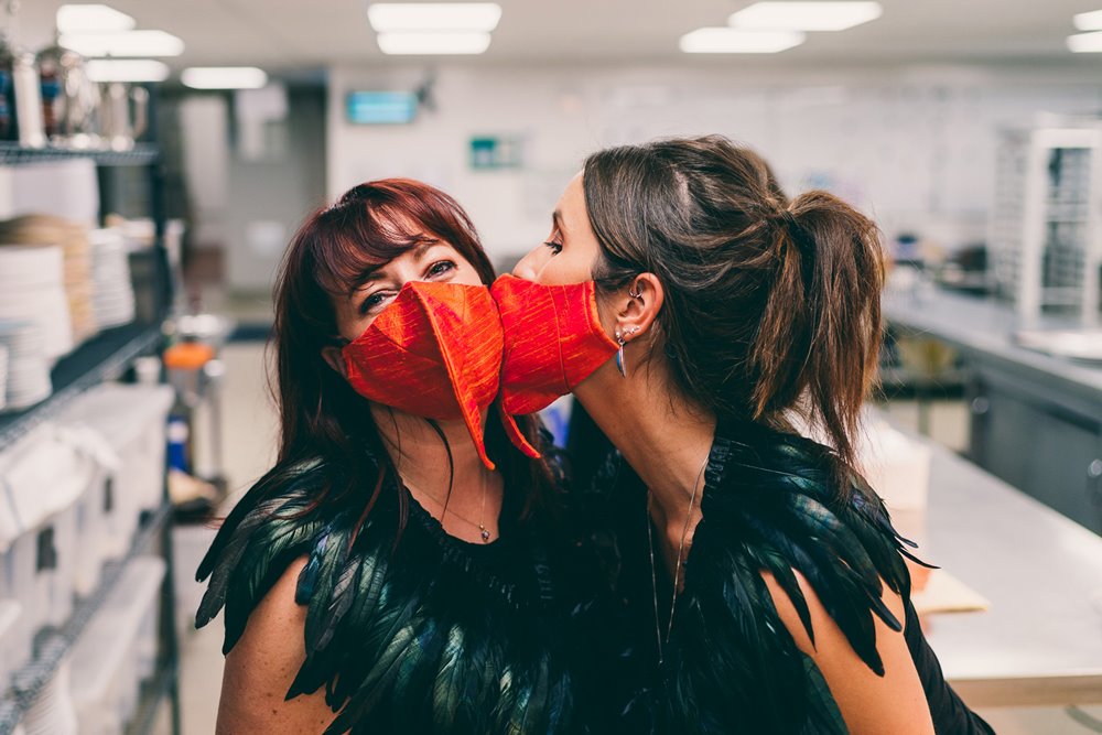 Beak to beak, two cabaret waitresses kiss on the cheek with covid beak masks