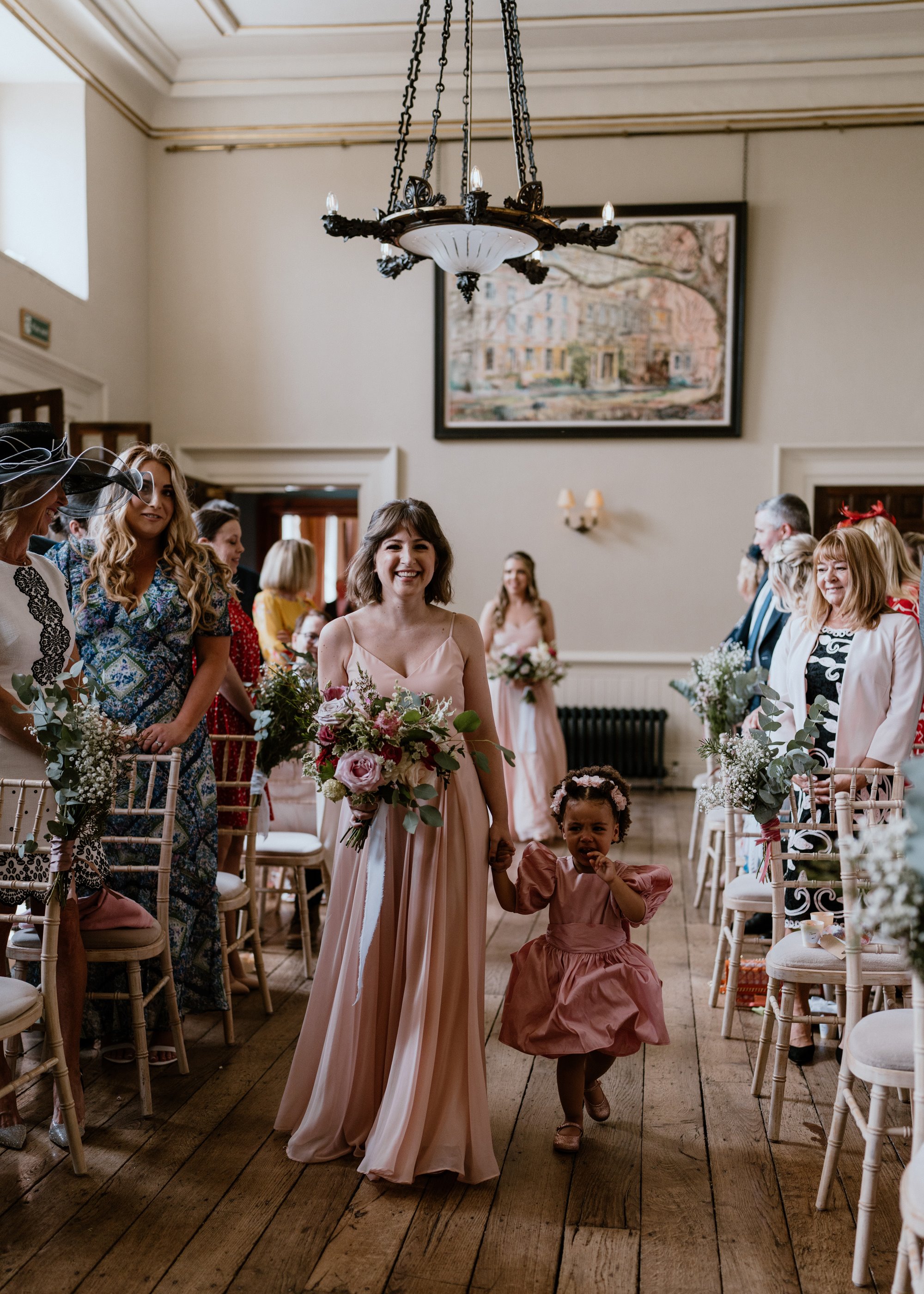 Bridesmaids and flower girl in pink dresses walk down the aisle in beautiful mansion house wedding venue