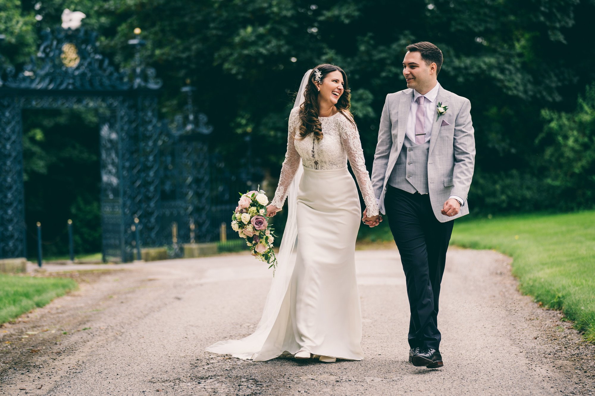 bride in pearl hair clip and lace dress holds hands with groom in grey suit walking up path to stately home wedding venue with wrought iron gates behind them