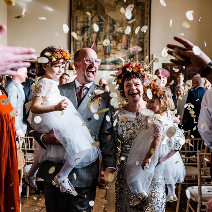 Fun creative couple bride in sequin dress and flower crown walk down the aisle with their children in arms with huge natural confetti in unique wedding ceremony in the uk