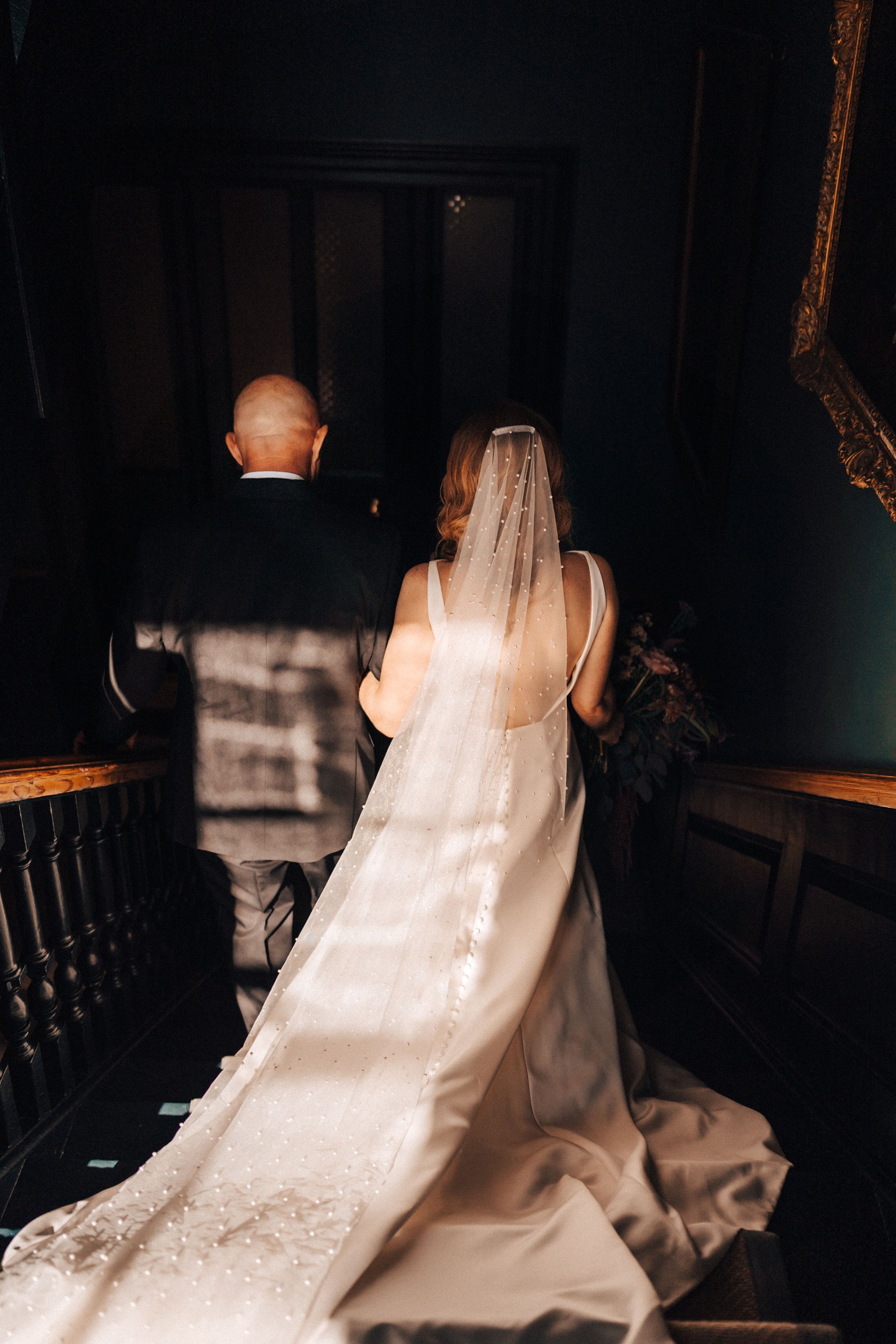 bride wearing long cathedral length veil with pearls walks with father of bride walks down the stairs of mansion house wedding venue to her ceremony