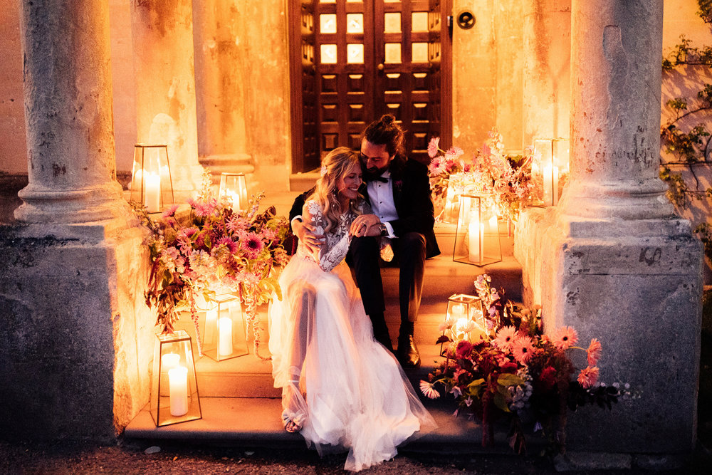 Cool bride and groom sit on steps of beautiful mansion house wedding venue surrounded by flowers and candles